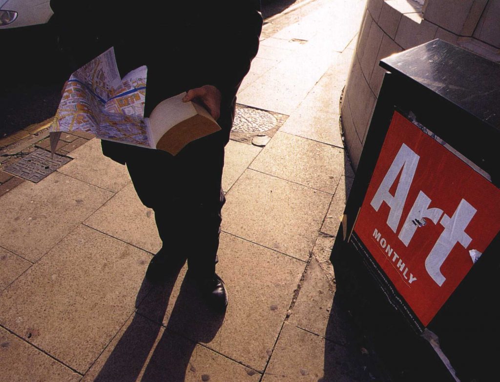 a person with a map and book on a street with an Art Monthly box in the foregound
